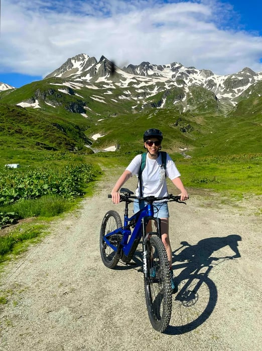 A woman stands beside a bike in a picturesque alpine meadow.