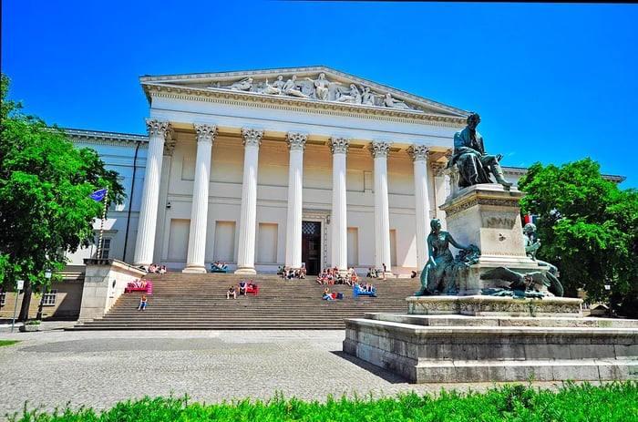 The facade of the Hungarian National Museum in Budapest showcases a grand white colonnaded building with visitors relaxing on its broad steps.