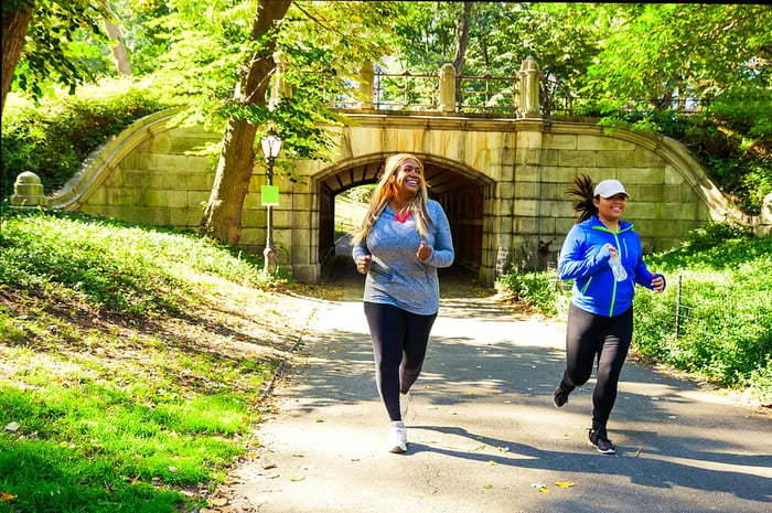 Two women run along a path in Central Park, smiling as they emerge from a tunnel beneath a decorative bridge.