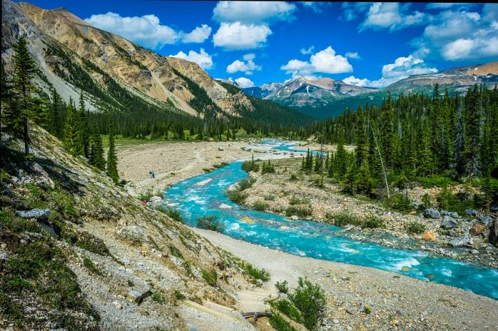 A flowing river meanders through a valley; some hikers follow a trail adjacent to it.