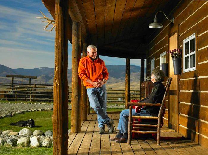 A senior couple beams on the porch of a rustic wooden cabin.