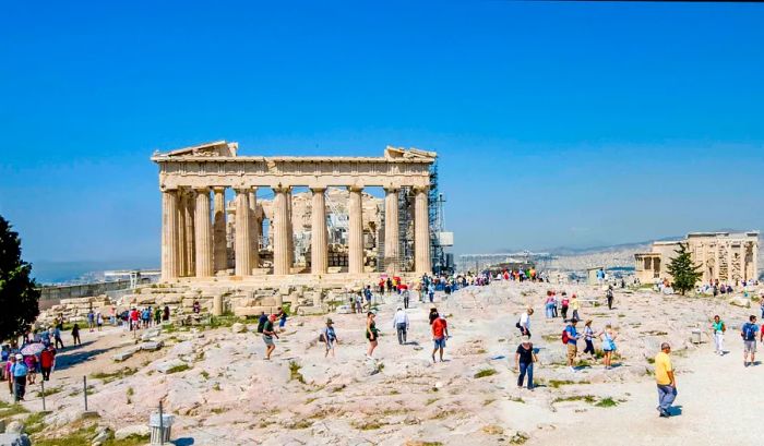 Visitors wander paths through an archaeological site on a sunny day, with the ruins of a columned temple in the background.