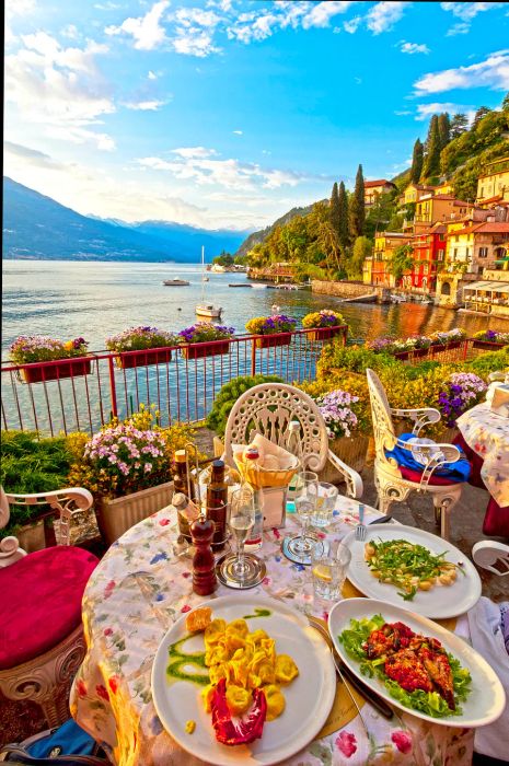 A trio of beautiful vegetarian Italian dishes served on elegant white plates at a charming outdoor terrace overlooking a serene lake, framed by historic buildings and alpine mountains in the distance.