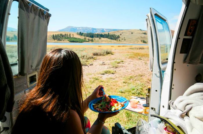 A woman enjoying a meal inside a van, gazing out at the stunning Yellowstone scenery