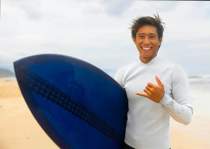 A happy man stands on the beach with a surfboard, beaming while posing for a photo.