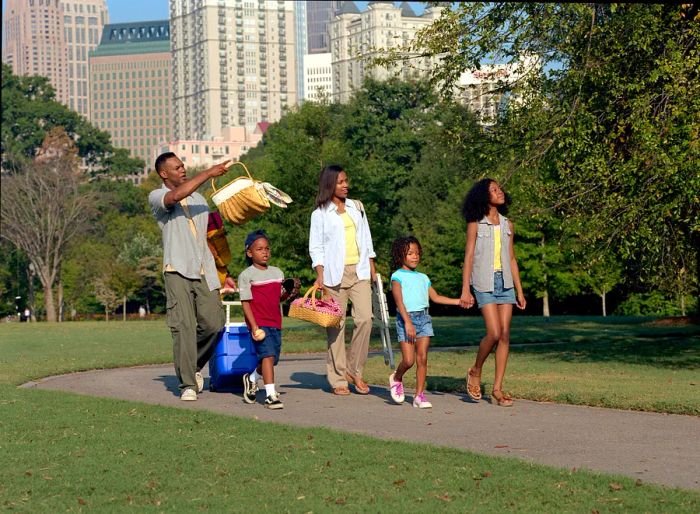 A family of five strolls through Piedmont Park in Atlanta, carrying picnic baskets and a cooler on wheels.