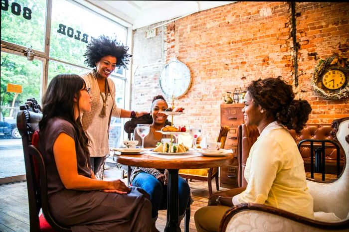 Three women are gathered around a table in a tea parlor, while another woman stands nearby, chatting with them