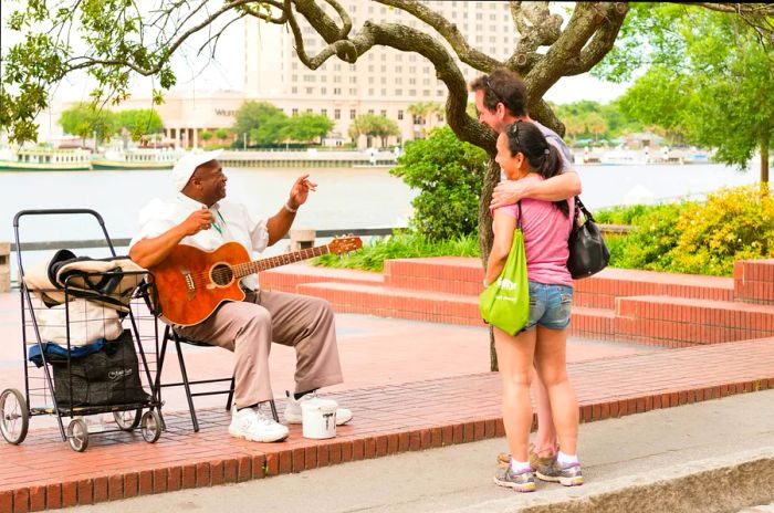 A couple stands together with their arms around each other, chatting with a street performer by the river