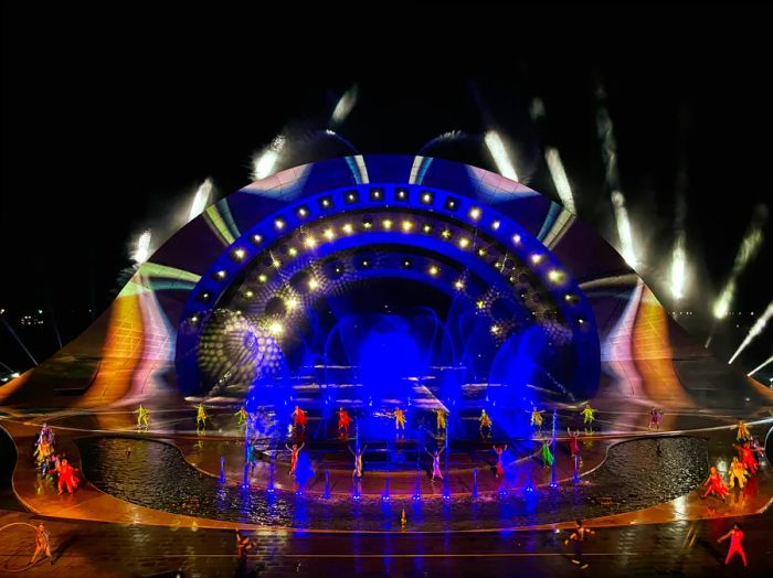 A group of dancers performs on a grand stage, illuminated by a spectacular light show and fountains.