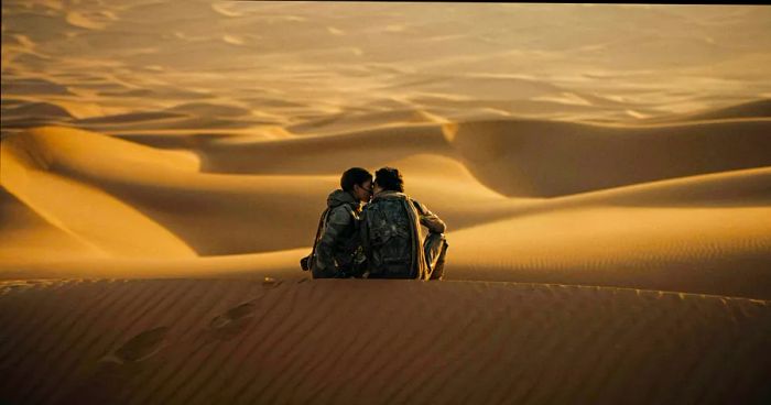 Two individuals perched on sand dunes in a vast desert lean in for a kiss