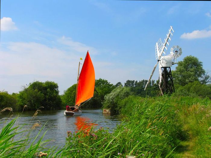 A sailboat glides past a windmill near The How Hill Trust and the River Ant in the Norfolk Broads, Ludlum, Norfolk.