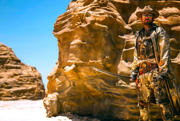 A man stands by a rocky outcrop, wielding a sword. The reflection of a group of people can be seen in his sunglasses.