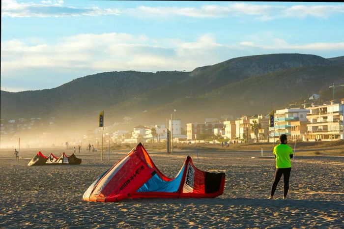 A young man in neoprene and lycra waits beside his kite on the sandy beach, with beachfront houses visible in the background, Castelldefels, Catalonia, Spain