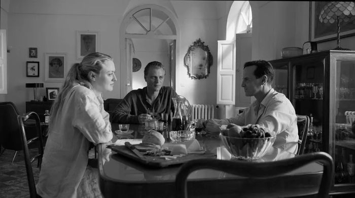 A black-and-white still from a film scene featuring two men and a woman sitting at a table, with the man and woman casting suspicious glances at the third man.