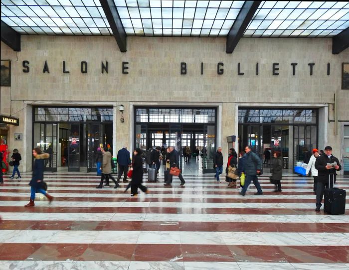 Passengers with luggage hurry past a station ticket office.