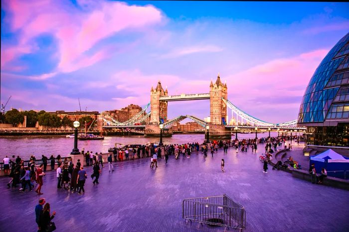 Crowds gather along the riverside near a grand Gothic bridge illuminated in the evening light.