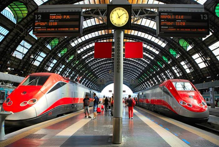 Two Frecciarossa trains are lined up, ready to depart from a grand city station with a stunning curved glass roof.