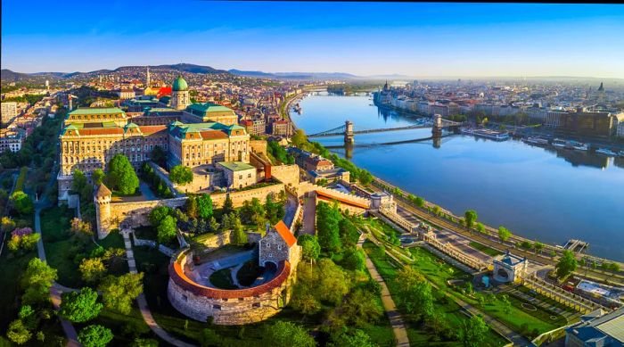 An aerial view of Buda Castle Royal Palace captured in the early morning, showcasing Széchenyi Chain Bridge, St. Stephen's Basilica, the Hungarian Parliament, and Matthias Church.