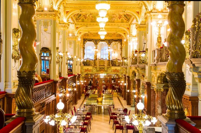 The lavishly decorated interior of the New York Café in Budapest features marble columns, opulent golden light fixtures, and luxurious crimson chairs.