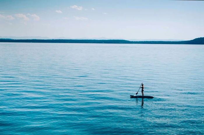 A lone figure balances on a paddleboard atop an exceptionally calm lake surface.