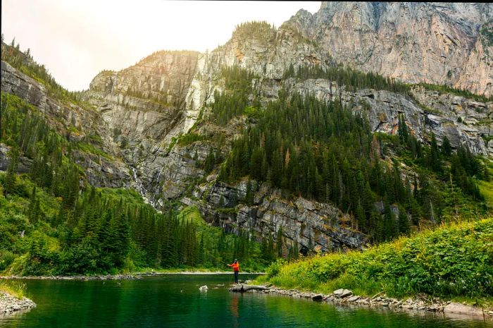 A fisherman casts a line into a river beneath the towering shadow of a massive granite cliff.