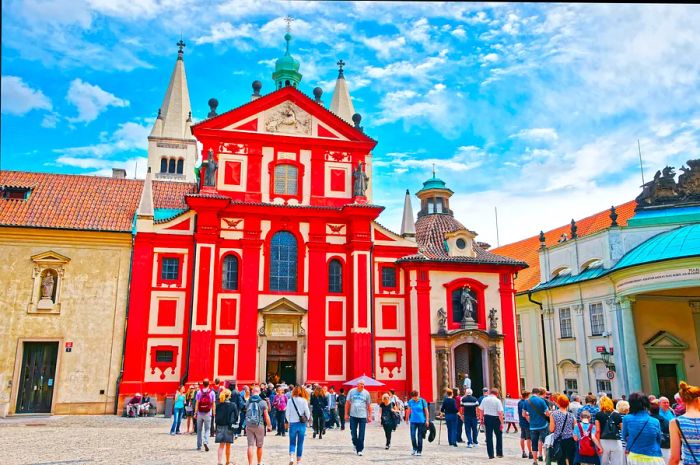 Tourists gathering around the vibrant red facade of St George Basilica at Prague Castle on a sunny June day.