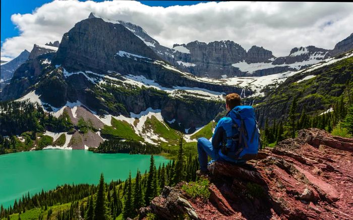 A hiker perches on a cliff, gazing down at an alpine lake.