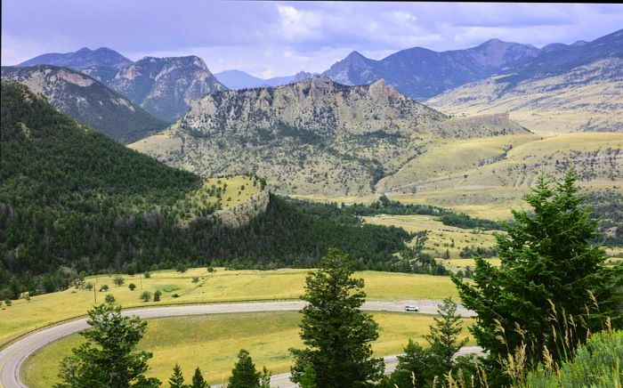 A car navigates a winding road through fields with mountains rising in the background.