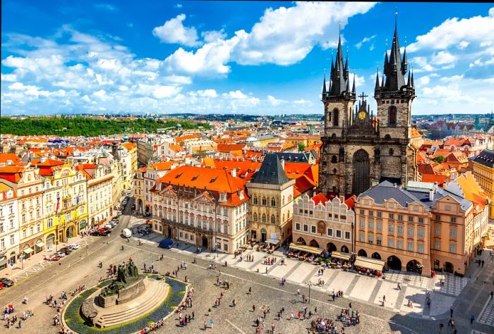 The towering twin spires of Tyn Church dominate Prague's Old Town Square, which buzzes with people strolling through and sitting at outdoor tables.