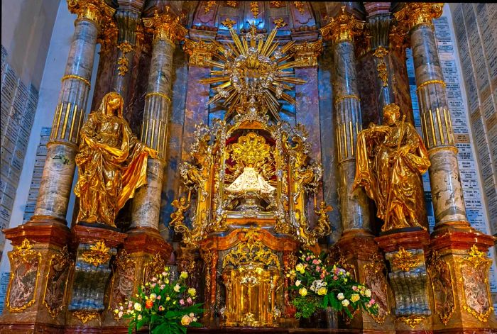 A small statue of the Infant Jesus is positioned at the center of an opulent, golden shrine within a Prague church. The statue is dressed in a white cloak.