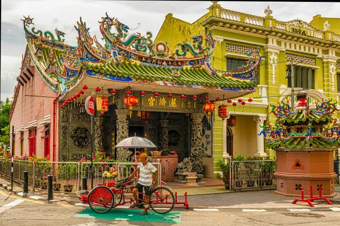 A man stands beside a rickshaw in front of a temple adorned with Chinese-style red lanterns.