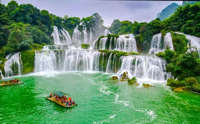 A multi-tiered waterfall with a bamboo raft filled with tourists enjoying the pool below