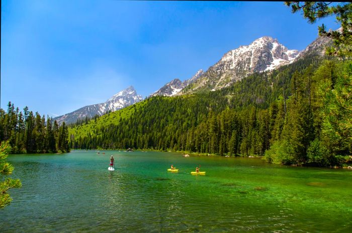 Paddleboarders and kayakers glide across crystal-clear waters at the foot of a mountain in Grand Teton National Park