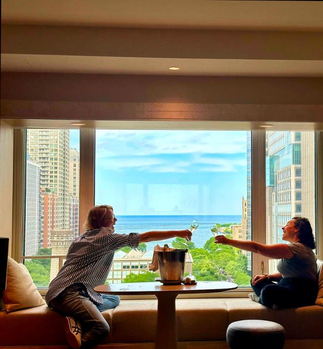 Two friends clink their wine glasses by a window overlooking the lake