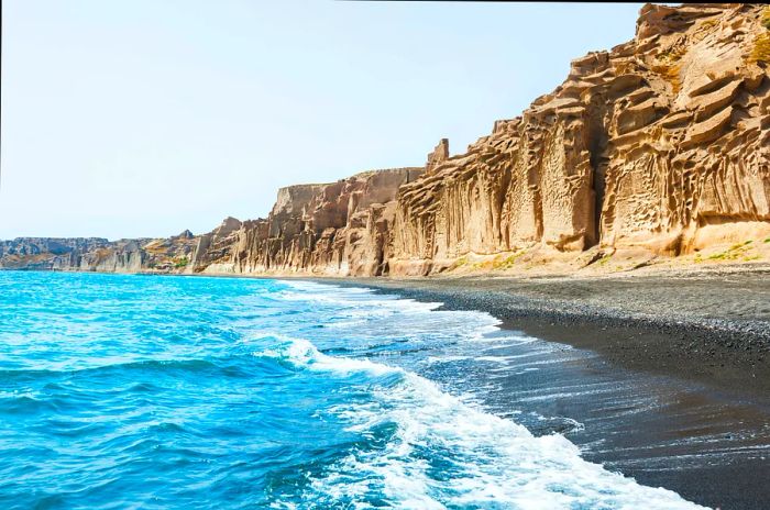 Waves gently caress the shores of a black-sand beach, with towering sandstone cliffs rising above.