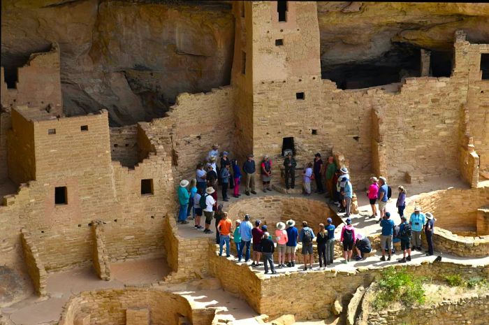 A group of tourists gathers in a circle at an ancient cliffside site, listening intently to their guide.