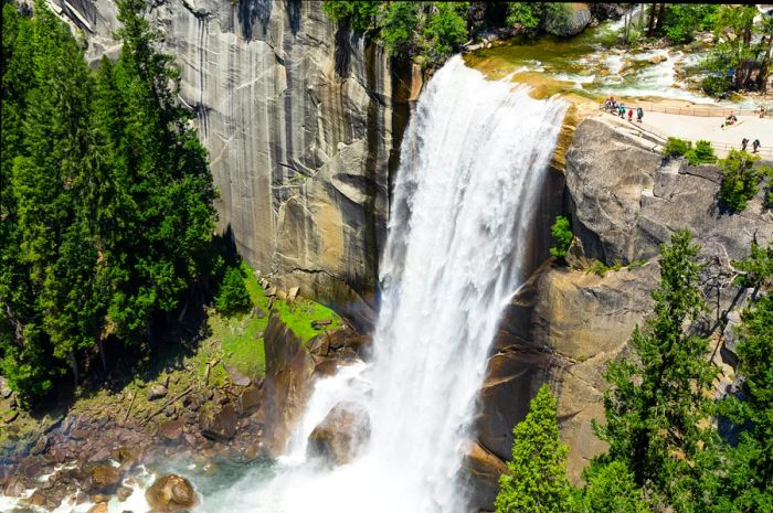 A powerful waterfall plunges into a shimmering pool below, while onlookers admire the view from a nearby platform.