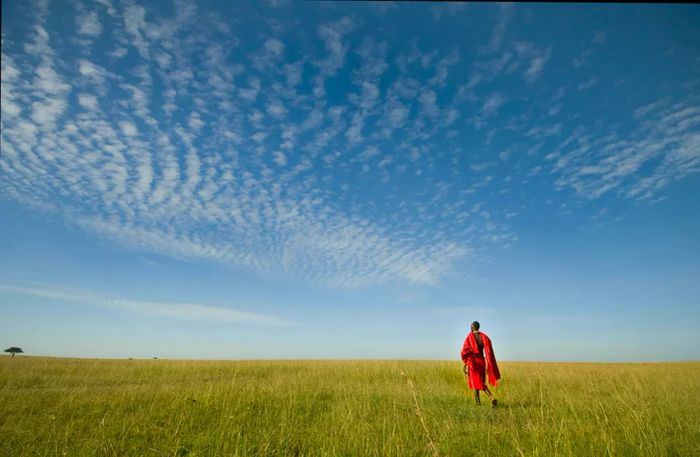 A solitary figure draped in a red checked tribal shawl strides across an open plain beneath the expansive sky.