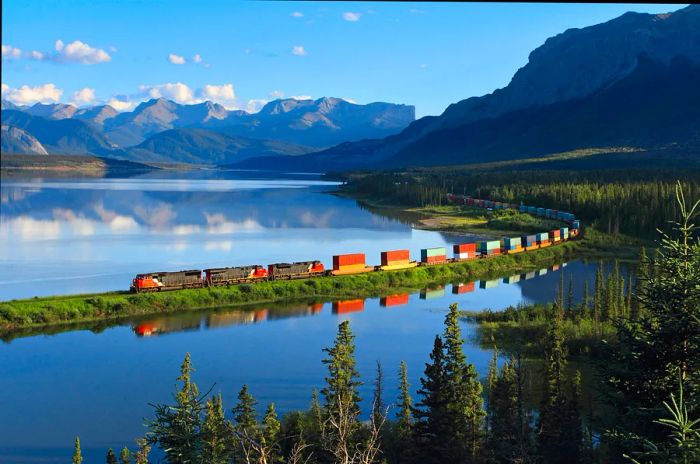 A lengthy freight train travels through breathtaking lakeside and mountain scenery