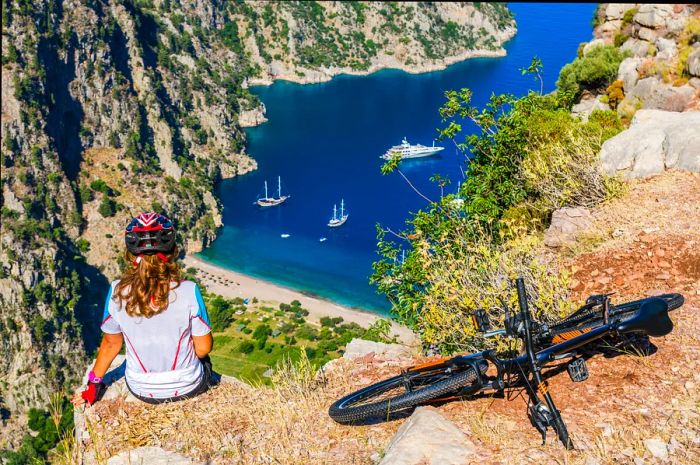 A woman relaxes beside her bicycle on a cliff overlooking deep blue waters dotted with boats.