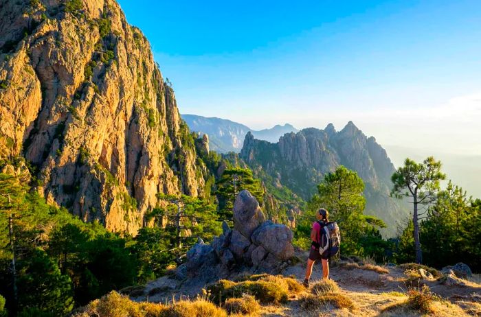 A woman with a backpack stands atop a hill along the GR20 trail, with rugged mountain peaks visible in the distance.