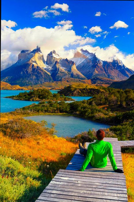 A hiker relaxes on a boardwalk overlooking a lake surrounded by majestic mountain peaks in Torres del Paine National Park.