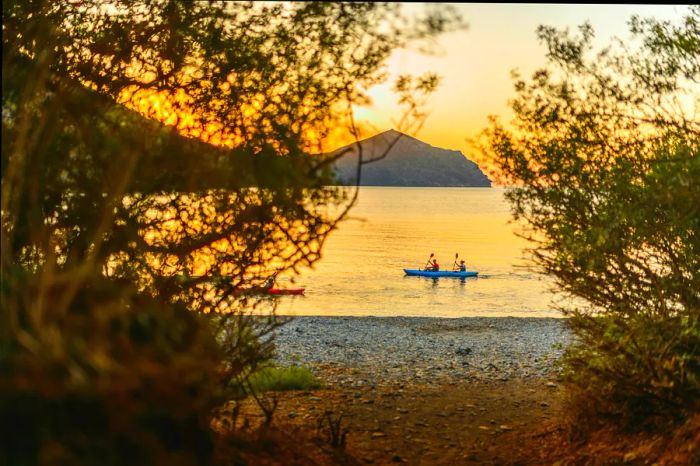 A distant view of friends kayaking along the Mediterranean shore as the sun rises over the headland in the background.