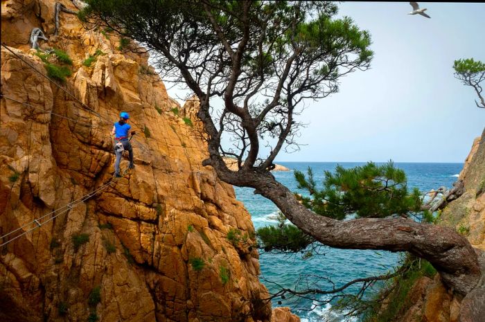 A woman climbs a via ferrata on a copper rock face overlooking the ocean in Costa Brava, Spain.