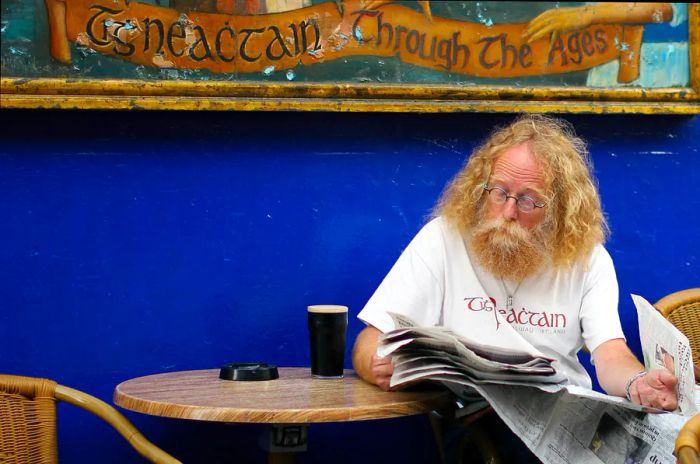 A man with long hair enjoys a pint of Guinness outside a vibrant blue pub.