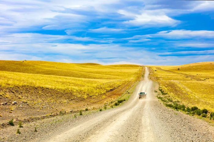 A vehicle travels along a secluded gravel road winding through expansive grasslands.