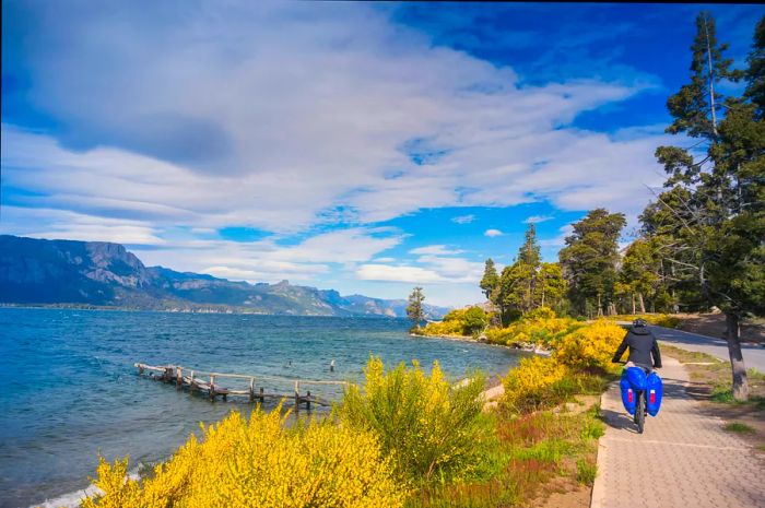 A cyclist rides beside a lake in a mountainous area, with bags attached to the bike