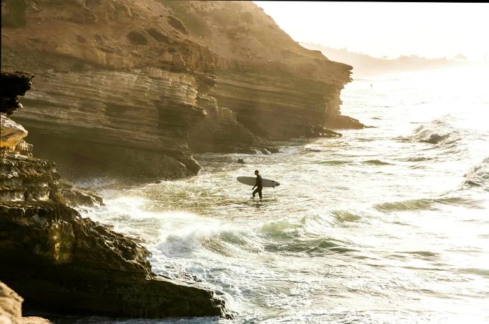 A surfer strides toward the rugged coastline at dawn, Taghazout Bay, Morocco