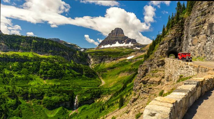 A vintage red car drives along a mountainous road in Glacier National Park, surrounded by lush green peaks.