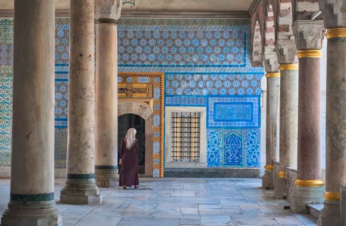 A woman approaches a doorway framed by intricately patterned blue tiles inside Topkapı Palace, which features several tall marble columns.